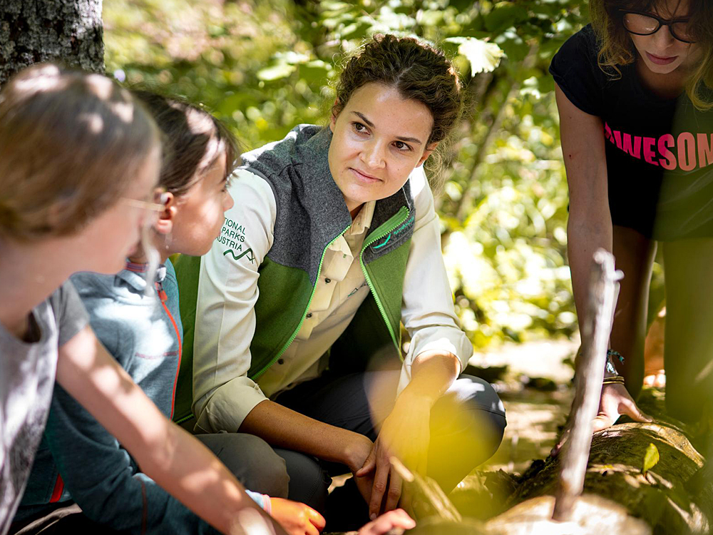 **Alt-Text:** Das Bild zeigt eine Nationalpark-Rangerin in grüner Dienstkleidung, die sich aufmerksam einer Gruppe von Kindern im Wald zuwendet. Gemeinsam sitzen sie am Waldboden und betrachten einen umgestürzten Baumstamm – eine Szene aus einem Naturbildungsprogramm im Nationalpark.