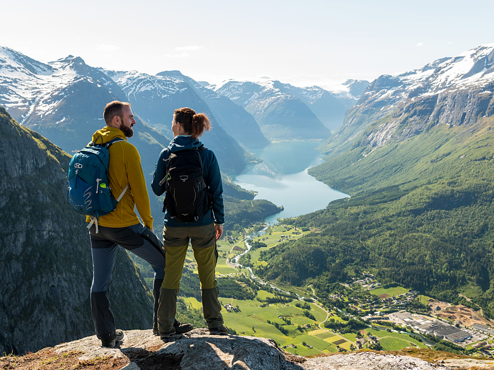 Das Bild zeigt zwei Wanderer – einen Mann und eine Frau – die auf einem Felsvorsprung stehen und in ein spektakuläres Tal mit einem langen, ruhigen See blicken. Umgeben sind sie von hohen, schneebedeckten Bergen und dichten Wäldern. Unter ihnen erstreckt sich eine grüne Landschaft mit Feldern, kleinen Straßen und Gebäuden. Die Szene wirkt friedlich und majestätisch – ein beeindruckender Blick in die Weite der Natur.