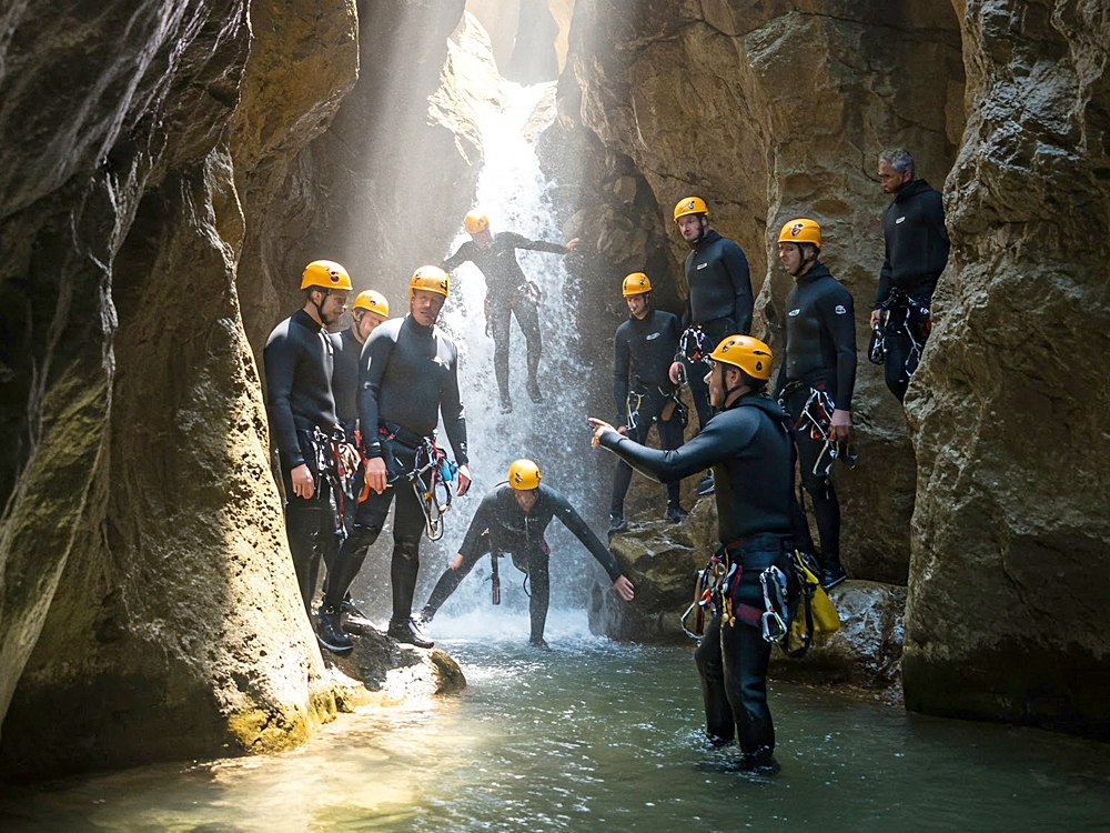 Das Bild zeigt eine Gruppe von Menschen beim Canyoning in einer felsigen Schlucht. Sie tragen Neoprenanzüge, Helme und Klettergurte. Ein Guide weist den Weg, während eine Person unter einem Wasserfall ins Wasser steigt. Sonnenlicht fällt dramatisch durch die Felsen.